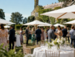 Outdoor summer wedding reception with guests mingling under parasols at a country house venue in hot weather.